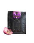 A black package of "Rest, Relaxation & Sleep Sampler Set" by Magic Hour with the label "Lucid Dreams" and a celestial design. A purple tag is attached at the top. A single pink flower with a yellow center rests beside the package. The packaging has gold accents and a mystical theme, perfect for loose leaf tea enthusiasts.
