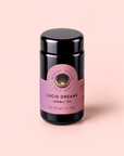 A black jar of Magic Hour Lucid Dreams: Tulsi-Turmeric Herbal Tea for Sleep & Calm with a purple label sits on a light pink background. The label displays the product name, weight (1oz/28g), and decorative gold accents, highlighting this soothing, caffeine-free chai infused with restful botanicals to support sleep and calm.