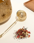 A golden tea infuser lies next to a small pile of Organic Tea leaves and flowers. An intricately designed golden container with star cutouts stands on the left, and a wooden tray is positioned in the background to the right, adding a touch of elegance to your Bestsellers of Magic Tea Starter Set- 60-75 cup sampler box by Magic Hour ritual.