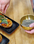 A person sprinkles Matcha Herbed Salt from a brass bowl onto toast with cherry tomatoes, greens, and fresh toppings on a black rectangular plate resting on a wooden surface.