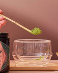 A hand holds the Ceremonial Matcha Bamboo Scoop filled with vibrant green matcha powder above a clear double-walled glass cup. A matching matcha jar and bowl, each holding more powder, rest on a wooden tray, capturing the serene atmosphere of a modern tea ceremony against a neutral backdrop.