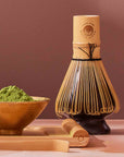 The Artisanal Magic Hour Matcha Whisk stands upright beside a bowl filled with vibrant green matcha powder and a bamboo scoop, all arranged on a light surface against a muted brown background, emphasizing the sustainable appeal of these handmade tea tools.
