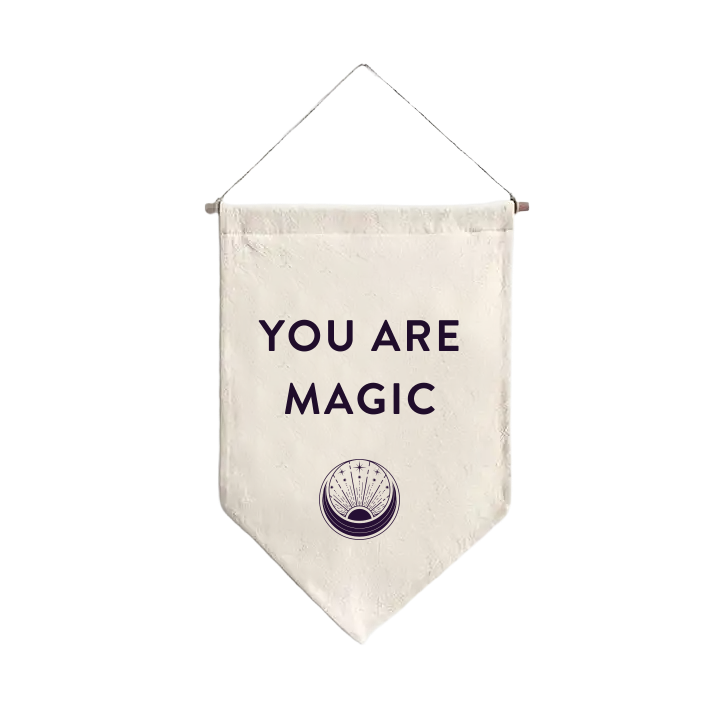 The You are Magic Flag is a cream-colored pennant made of natural fabric, featuring the bold, black phrase "YOU ARE MAGIC." Beneath this empowering text is a mystical circular emblem. The flag hangs elegantly from a thin thread attached to a wooden rod.