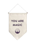 The You are Magic Flag is a cream-colored pennant made of natural fabric, featuring the bold, black phrase "YOU ARE MAGIC." Beneath this empowering text is a mystical circular emblem. The flag hangs elegantly from a thin thread attached to a wooden rod.
