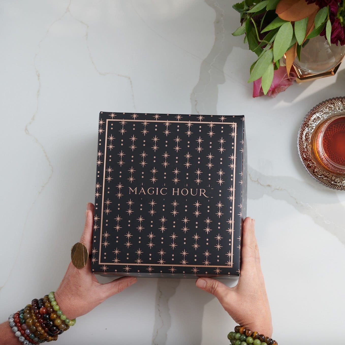 Two hands holding The Lovers Box, which is a black box adorned with gold star patterns and the text "MAGIC HOUR Branded Gift Box." The person wearing multiple beaded bracelets and a large ring. A cup of Lovers tea blend, crafted from women’s balancing herbs, and a floral arrangement are placed on the right side of the white marble surface.