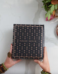 Two hands holding The Lovers Box, which is a black box adorned with gold star patterns and the text "MAGIC HOUR Branded Gift Box." The person wearing multiple beaded bracelets and a large ring. A cup of Lovers tea blend, crafted from women’s balancing herbs, and a floral arrangement are placed on the right side of the white marble surface.