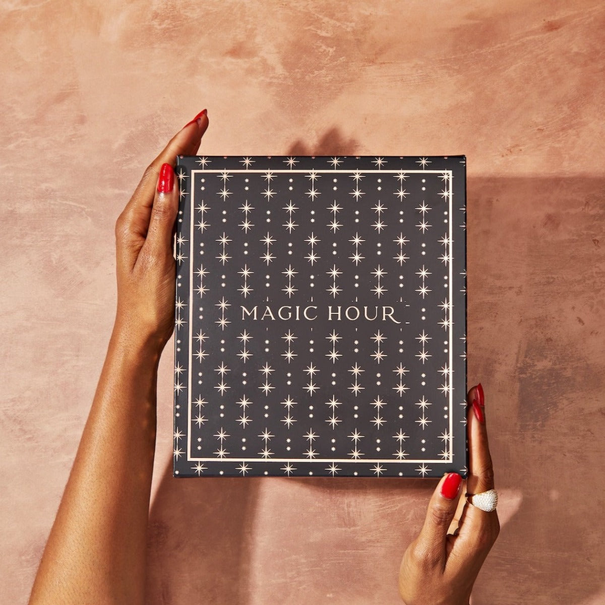 A pair of hands with red nail polish and a ring holds The High Priestess Box, which is black and adorned with a pattern of small, white, star-like symbols. "MAGIC HOUR" is written in white text at the center. The background is a textured, rose-colored surface.
