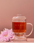 A Glass Tea Infuser, elegantly designed and currently filled with tea, rests on a light wooden surface against a muted pink background. A pink chrysanthemum flower lies beside the infuser, adding a delicate touch to the composition.