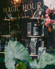 A storefront for Ojai Tea Salon displays "MAGIC HOUR" and "TIME FOR INFINITY" in gold. Reflected leafy plants frame tea essentials and the Tea Essentials + In Person & Virtual Tea School Course, all visible through the glass.