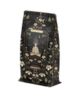 A black pouch with an intricate design contains Magic Hour's "The Oracle: Lemon Lavender Tea for Supporting the Gut-Brain Axis Case Packs." This caffeine-free blend features a meditating figure surrounded by flowers, celestial symbols, and ornate patterns. Infused with adaptogenic roots, the packaging also showcases white floral and vine elements in the background.