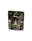 A dark, intricately designed bag features a central illustration of a meditating figure surrounded by celestial elements like stars, moons, and swirling patterns. Adorned with blooming flowers and leaves in a gold and green palette, it holds The Oracle: Lemon Lavender Tea for Supporting the Gut-Brain Axis Case Packs. The label reads "MAGIC HOUR THE ORACLE.