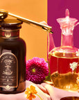 A dark jar labeled “Magic Hour” is paired with the Perfect Cup Tea Scoop with Pouch Clip in gleaming gold, resting beside a glass decanter of liquid and surrounded by colorful flowers on a pink and orange background—an elegant scene showcasing exquisite tea accessories.