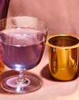 A clear glass of water stands next to the Midas Touch: Golden-Hued Tea Strainer cup on a textured, light pink surface with a peach-colored flower nearby.