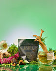 A packet labeled "Earth: Stabilizing and Nourishing Herbs, Roots, and Puerh tea" rests among moss, flowers, and a wooden stick, with a glass cup of grounding tea in front. The green gradient background enhances the calming atmosphere.