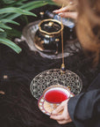 A person holds a cup of red tea beside the Crystal Grid: Flower of Life Tea Ceremony Altar, with greenery in the background. A crystal pendulum is suspended above the grid, evoking a ritual or meditation focused on intentional manifestation.