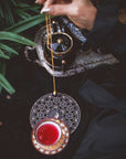 A hand holds a pendulum above the Crystal Grid: Flower of Life Tea Ceremony Altar, surrounded by red tea in an ornate cup, a teapot, and green leaves, evoking a mystical, tarot-inspired setting for ritual tea ceremonies.