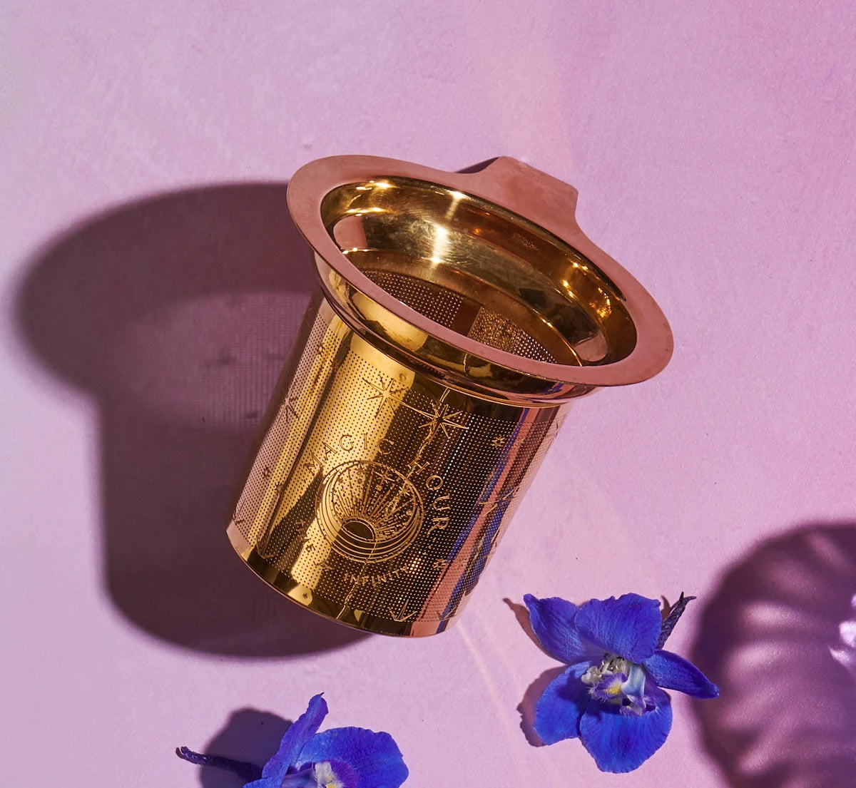 Product Name: Loose Leaf Tea Starter Set
A gold metal tea infuser from the Loose Leaf Tea Starter Set, featuring an intricate mesh design, rests on a pink surface and casts a shadow. Nearby, two blue flowers enhance the scene with their vibrant color.