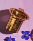 Product Name: Loose Leaf Tea Starter Set
A gold metal tea infuser from the Loose Leaf Tea Starter Set, featuring an intricate mesh design, rests on a pink surface and casts a shadow. Nearby, two blue flowers enhance the scene with their vibrant color.