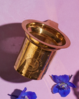 A gold-toned Steep The Stars - Artisan Tea Strainer by Magic Hour with intricate patterns is placed on a pink surface. Next to it, a small, vibrant blue flower petal adds a touch of color. The shadow of the Steep The Stars - Artisan Tea Strainer is cast on the surface, enhancing the image's depth—a perfect setting for brewing your favorite loose leaf tea.