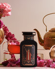 A black bottle labeled "The Magician," evoking transformative abilities, is surrounded by pink flowers, a glass cup of tea, a golden teapot, and a glass orb. The background features more pink flowers and a light-colored backdrop, creating a luxurious and elegant setting with wellness benefits.