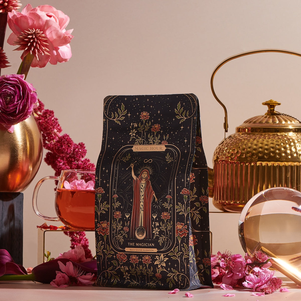 A black tea package from "The Magician Box" featuring an intricate design is elegantly displayed alongside a gold teapot, a glass cup of tea, pink flowers, and a Luxe Loose Leaf Tea Strainer, all artfully arranged on a tabletop.