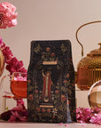 A black tea package from "The Magician Box" featuring an intricate design is elegantly displayed alongside a gold teapot, a glass cup of tea, pink flowers, and a Luxe Loose Leaf Tea Strainer, all artfully arranged on a tabletop.