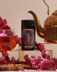 A delicately arranged scene features a teapot with a hammered texture, a glass cup filled with red tea, a dark jar labeled "The Magician Box," pink flowers, and a decorative gold tray with dried flower petals and an elegant Luxe Loose Leaf Tea Strainer, all set against a neutral background.