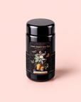 A beautifully designed canister of Mango Jasmine Oolong Iced Tea sits on a light pink background, featuring floral and fruit illustrations, “Cosmic Garden Iced Teas Mango Jasmine” on the label, and “Magic Hour” at the bottom. This eye-catching tin captures the essence of metabolism-boosting iced tea, delivering vibrant mango jasmine oolong tea flavor with every refreshing sip.
