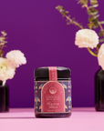 A decorative jar of "Ceremonial Bath Salt Soaks" in a violet glass jar rests on a pink surface, with blurred white flowers and vases against a purple backdrop—an elegant addition to your next self-care ritual.