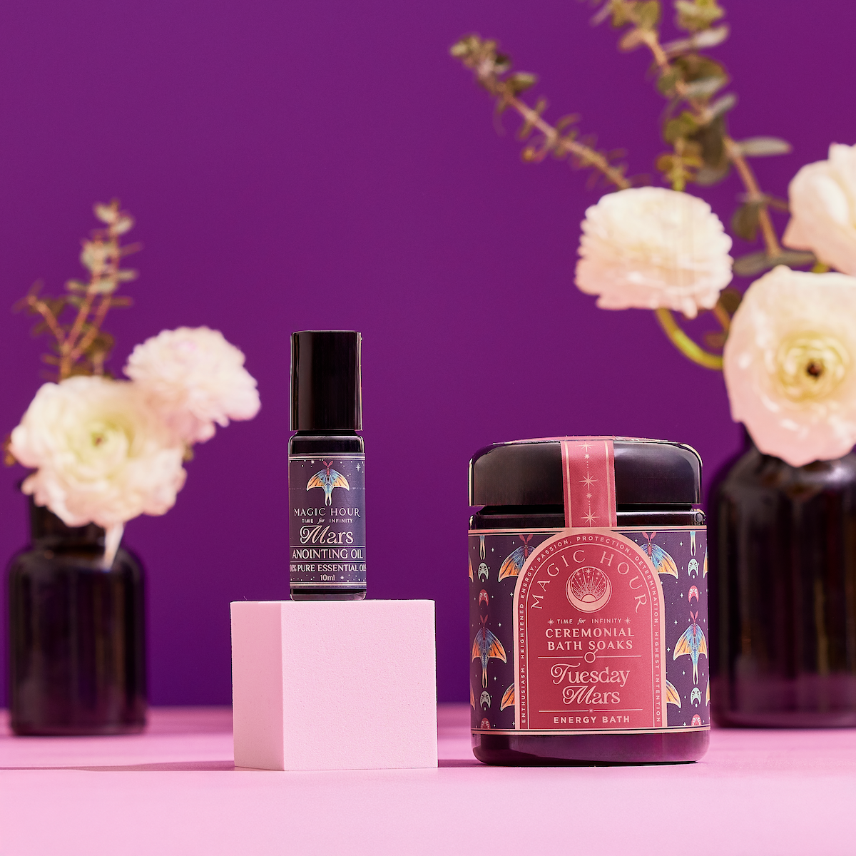 A bottle of anointing oil and a jar of bath soaks from The Celestial Self-Care Ritual Set are displayed on a pink surface, accented by white flowers in dark vases, all set against a vivid purple background.