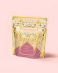 A pastel tea package labeled "Magic Hour" stands upright, featuring gold, green, and purple Art Deco designs. The label reads "Pumpkin Spice Marzipan White Tea." Set against a soft pink background, it has a decorative flair.