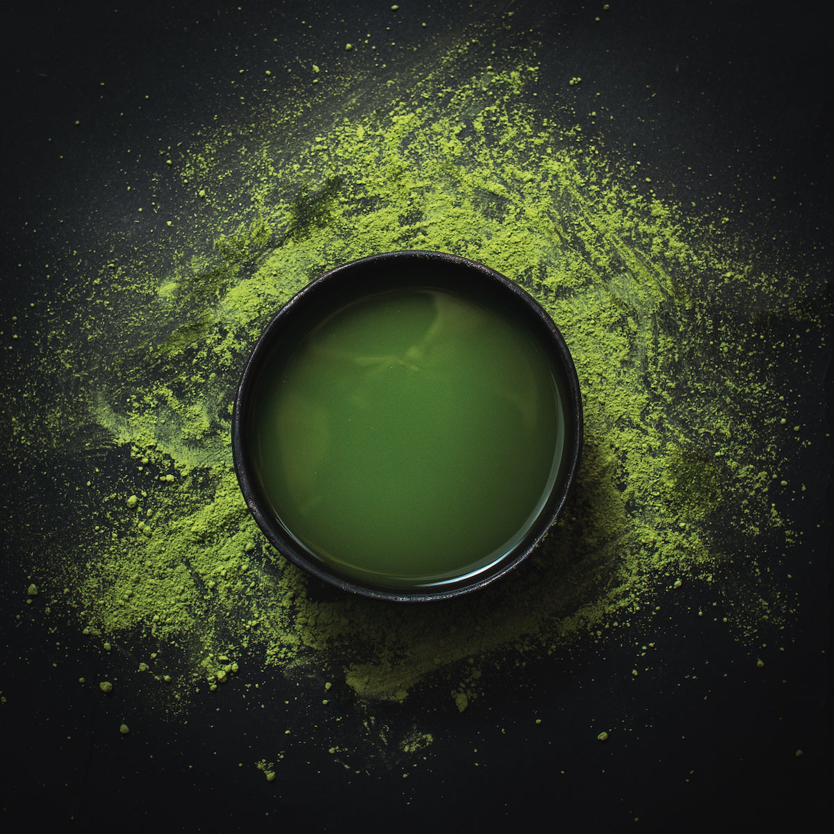 A cup filled with vibrant green matcha tea is placed on a dark surface. Green matcha powder is scattered around the cup, creating a striking contrast against the dark background. The overhead lighting emphasizes the rich color and texture of the matcha powder.