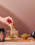 A bamboo whisk stirs Organic Ceremonial Matcha 700 in a glass bowl. On a wooden tray are a jar of matcha powder, a bowl with matcha, and a small black cup.