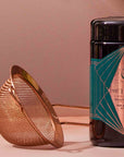 A Rose Gold Matcha Sift rests beside a black jar adorned with a turquoise and copper geometric label on a wooden surface, ready to elevate your matcha experience.
