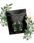 A black packet of Matcha Herbed Salt, adorned with golden stars and botanical designs, is displayed among green leaves, white flowers, and garlic bulbs—a vibrant showcase of this gourmet seasoning blend on a light background.