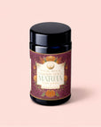 A black jar labeled “Pumpkin Spice Matcha” with festive orange pumpkins and foliage—evoking autumn botanicals—adorns a dark maroon and gold label, displayed against a pale pink background. This seasonal blend features matcha infused with pumpkin spice for a unique autumn twist.