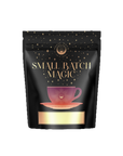 A black pouch labeled “Small Batch Magic” with gold stars, a gold sun emblem, and a purple and orange teacup illustration contains Cherry Cola Black Tea: Original Formula. A blank gold label is at the bottom.