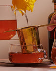 A hand is seen pouring tea through a Midas Touch: Golden-Hued Tea Strainer by PMB Mesh Screen Company into a glass teacup. A teapot filled with amber liquid is also visible. Another teacup is partially seen in the foreground. A daffodil in a glass container and a dark bottle are in the background.