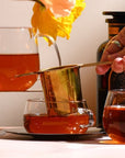 A hand holding an artisan-crafted gold tea strainer from the Magic Hour Starter Kit by Magic Hour is pouring tea into a glass cup on a saucer. Another glass teacup and a small glass teapot containing amber tea are on the surface. Bright orange flowers and a dark amber bottle in the background exude the elegance of a traditional tea ceremony.