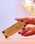 A hand with dark nail polish holds the Mindful Minutes Tea Timer box, labeled "MAGIC HOUR MINDFUL MINUTES," against a soft pink background with blurred white flowers, highlighting its elegant design for precision tea steeping.