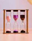 The Mindful Minutes Tea Timer features a wooden frame holding three hourglasses with pink, purple, and black sand, each partially emptied for precise tea steeping, set against a soft pink background.