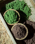 Two wooden bowls sit on a tray: one holds vibrant Mint Matcha Cocoa and the other, rich chocolate mint powder. Some powder is scattered nearby, and a woven mat is partially visible beside the tray.