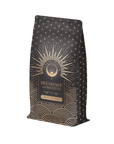 The "Mood Balance: Breakfast with Benefits" coffee bag in black and gold displays intricate geometric patterns and the text "Magic Hour" on a central design, enriched with ashwagandha root for a revitalizing touch.