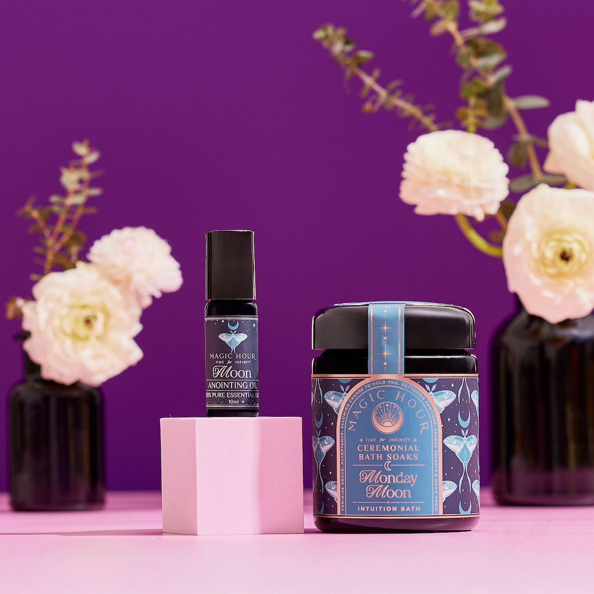 Two Magic Hour products—a small bottle and a jar labeled "Ceremonial Bath Soaks"—sit on a pink block with white flowers in black vases and a purple background, featured as The Celestial Self-Care Ritual Set for your perfect self-care experience.