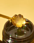 A Gold Floral Teaspoon drizzles golden, flower-shaped honey into an open jar, as bright light sparkles off the honey like stars against a yellow background.