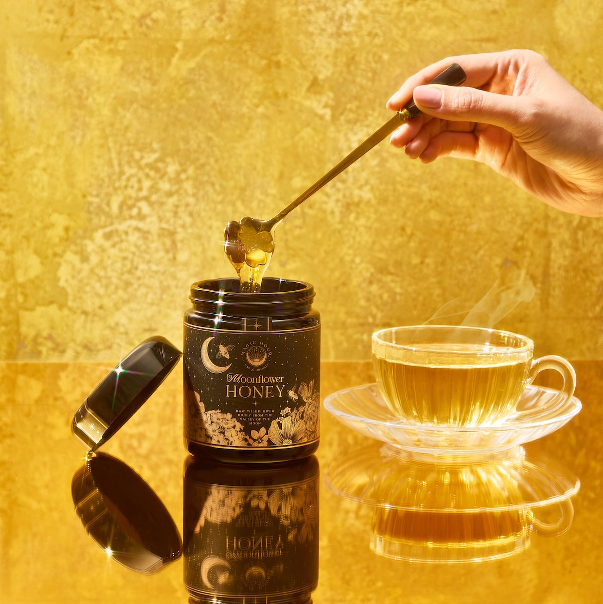 A hand drizzles Moonflower Honey—100% raw, antioxidant-rich wildflower honey from Ojai—into a teacup on a reflective golden surface, set against a gold-toned background that enhances the warm, luxurious atmosphere.