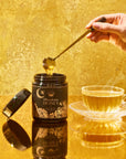 A hand drizzles Moonflower Honey—100% raw, antioxidant-rich wildflower honey from Ojai—into a teacup on a reflective golden surface, set against a gold-toned background that enhances the warm, luxurious atmosphere.