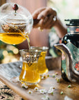 A hand pours Moonlight in Marrakesh: Spiced Mint Green Tea from a glass teapot into ornate glasses on a wooden tray, with flowers and a decorative black bottle nearby.