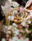 A person with manicured nails holds the lid of a glass teapot filled with Moonlight in Marrakesh: Spiced Mint Green Tea, surrounded by flowers and a dark glass bottle on a patterned surface.