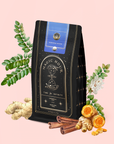 A black and gold tea bag labeled "Magic Hour" with an hourglass design and a blue "Moringa Moon" seal sits among ginger root, cinnamon sticks, turmeric, and green leaves, highlighting Moringa Moon Herbal Tea, a caffeine-free blend.